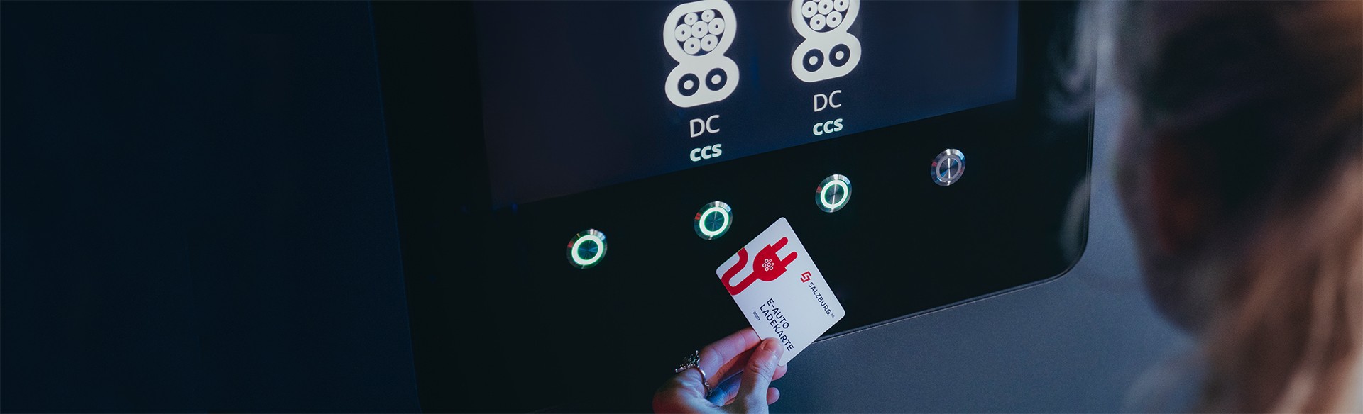 Hand hält Salzburg AG E-Auto Ladekarten vor Ladestation mit DC- und CCS-Anschlüssen.