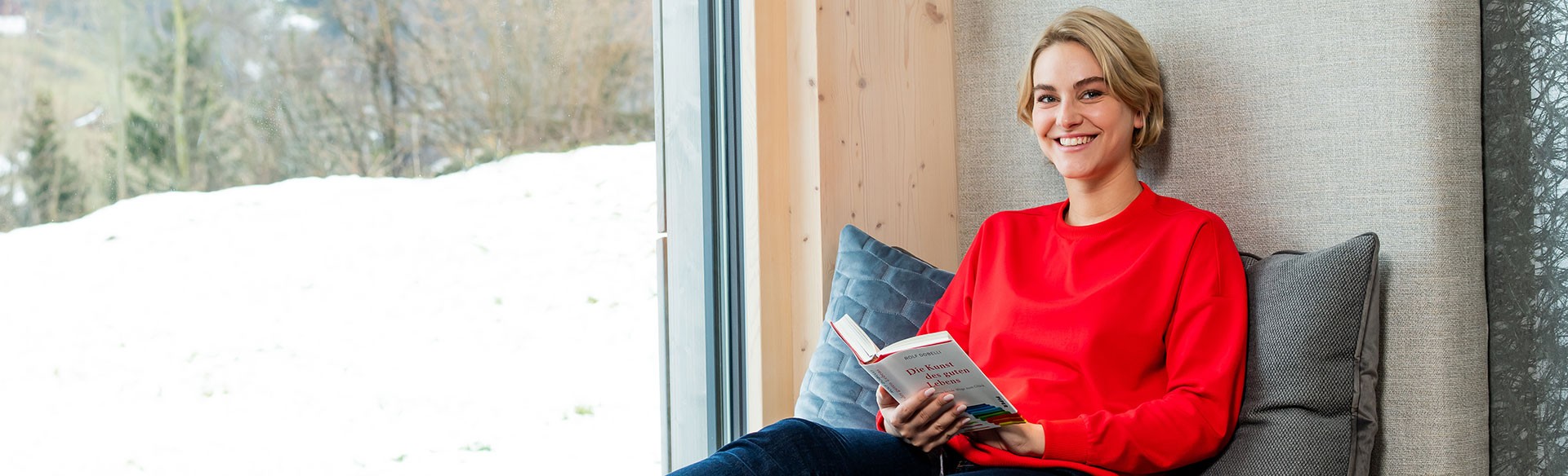 Frau liest Buch am Fenster mit Winterlandschaft im Hintergrund.